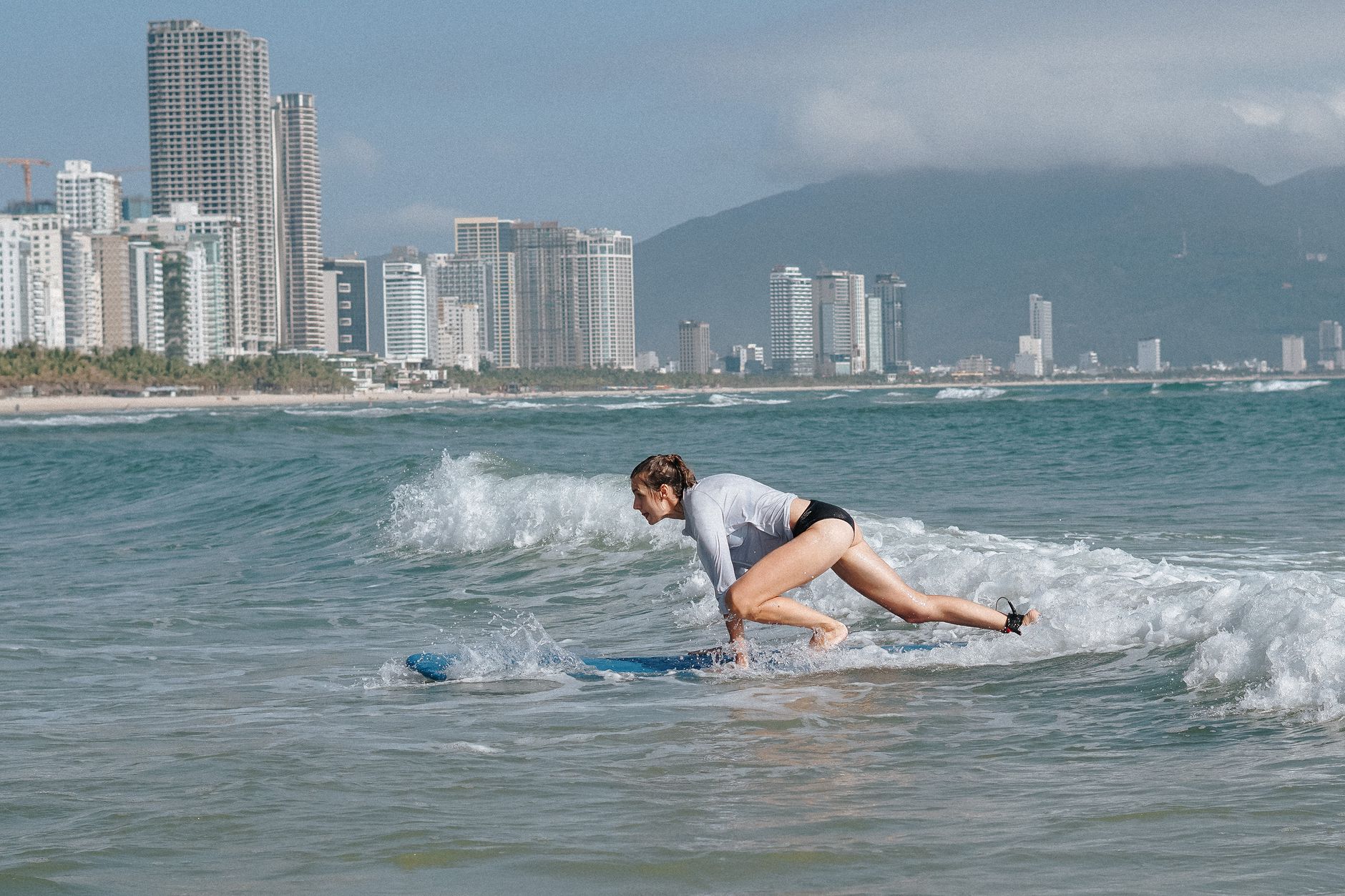 A woman surfing on a small wave near a city skyline with tall skyscrapers in view.